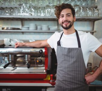 Portrait of smiling waiter standing with hand on hip at counter in cafÃ©