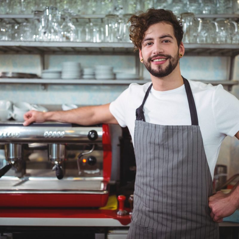 Portrait of smiling waiter standing with hand on hip at counter in cafÃ©